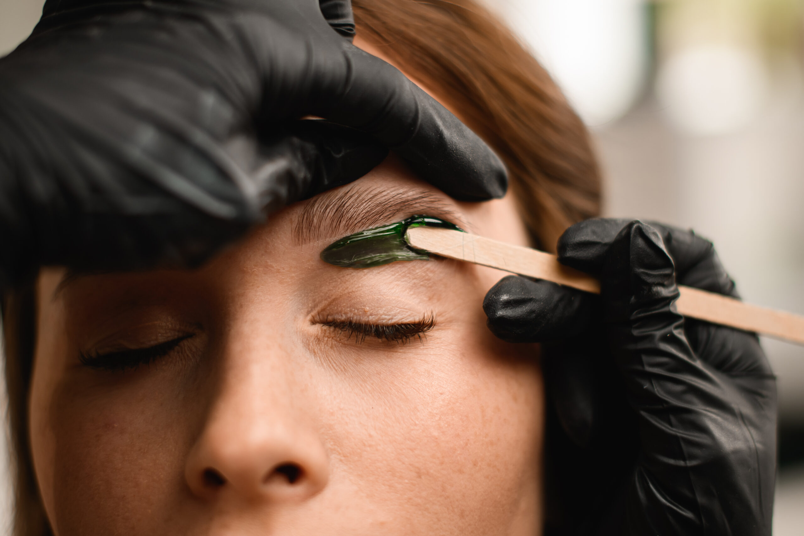 Close-up of the process of applying green wax to skin at female eyebrows to remove unwanted hair. Professional correction in beauty salon.